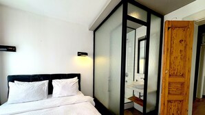 A bedroom with a bed, white linens, a nightstand, a wall-mounted light, and a wooden door leading to a bathroom with a sink and mirror. - Galata Port Flats Chakra 2 (Istanbul)
