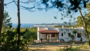 A white villa with a tiled roof, surrounded by trees and a view of the sea and distant hills. - Casa Para Familias en Ibiza (Sant Josep de sa Talaia)
