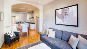 Un salon moderne avec un canapé gris, un fauteuil noir et une table basse. On y trouve également un coin repas avec une table et des chaises en bois, et une cuisine à l’arrière-plan.