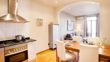 A modern kitchen with a stove, oven, and stainless steel range hood. There is a dining area with a wooden table, white chairs, and a sofa.