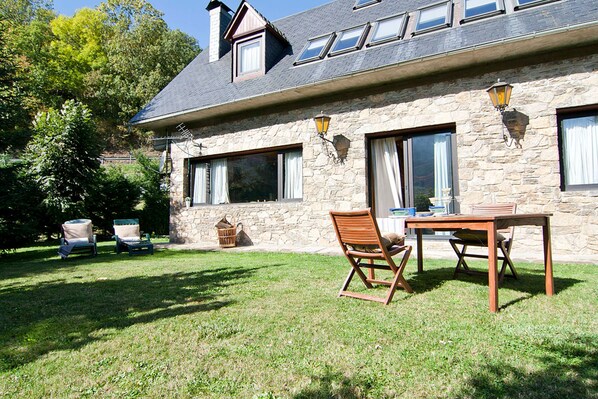 A stone building with a sloped roof, a dining table with chairs, and a grassy yard.