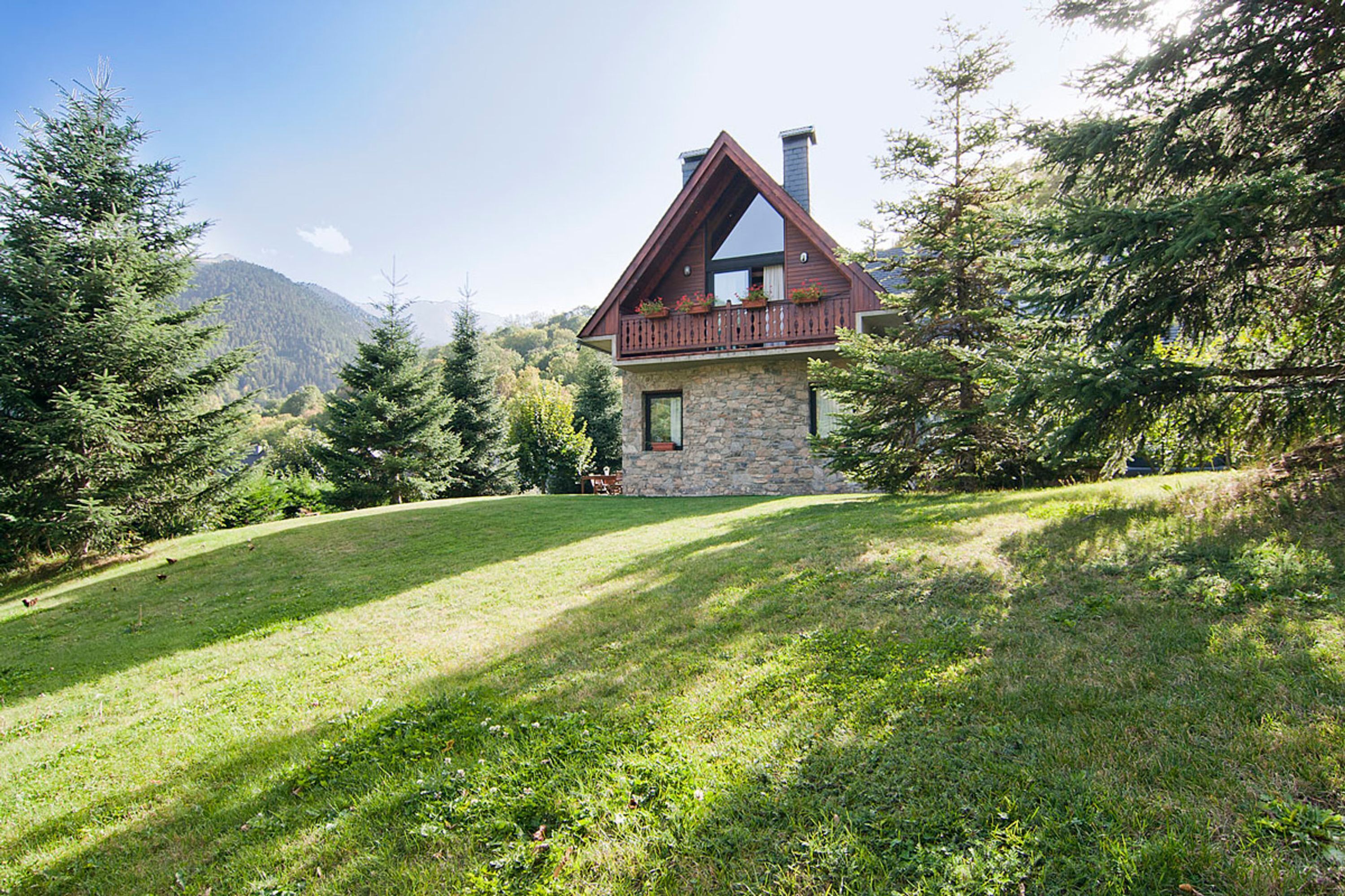 A stone house with a wooden balcony, surrounded by trees and a grassy hill.