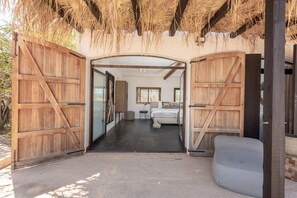 A room with a bed, a small table, and a chair. There are wooden doors and a thatched roof.