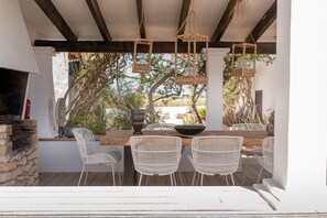 An outdoor dining area with a wooden table, white wicker chairs, hanging lanterns, and a stone wall grill.