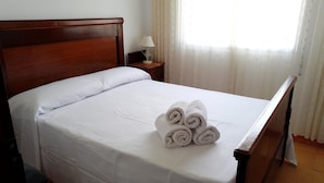 Un letto ben rifatto con lenzuola bianche e asciugamani arrotolati, una testiera in legno, un comodino con una lampada e una finestra con tende trasparenti.