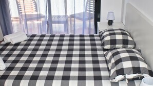 A hotel room with a checkered bedspread, a white pillow, a white towel, a lamp, and a view of outdoor chairs through a window. - Apartament Sergi in Amposta (Amposta)