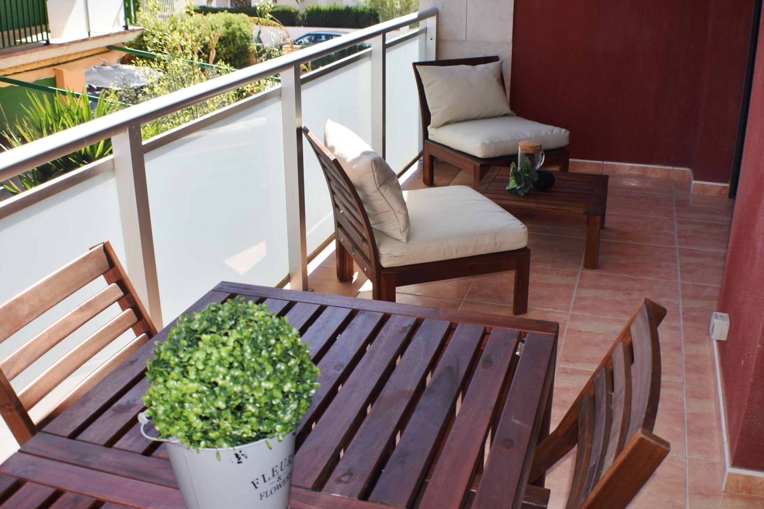 Una mesa de madera con una planta en maceta, una silla de madera y una silla con cojín y almohada en un balcón con barandilla.