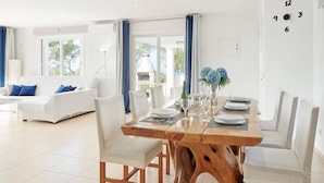A modern dining area with a wooden table, white chairs, and a white sofa in the background. - Villa Ses Abelles in Sant Josep de Sa Talaia (Sant Josep de sa Talaia)