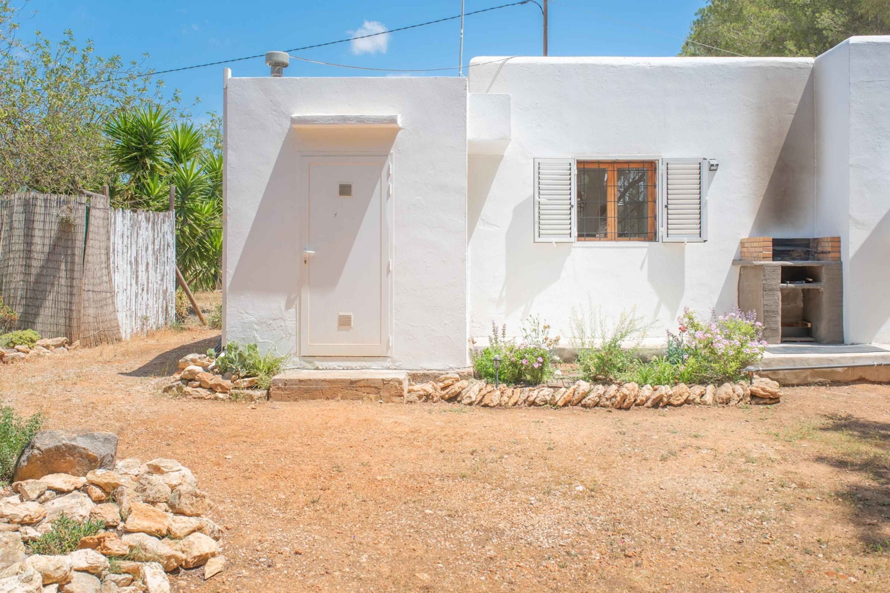 A white building with a door and window, surrounded by a dry landscape and a stone wall.