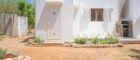 A white building with a door and window, surrounded by a dry landscape and a stone wall.