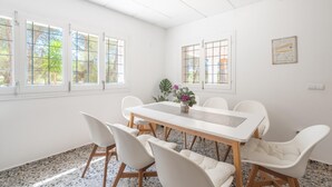 A bright, spacious room with a large table and white chairs, large windows with a view of greenery, and a framed picture on the wall. - Can Vert - Casa Rural Ideal Para Desconectar (Santa Eulalia del Rio)