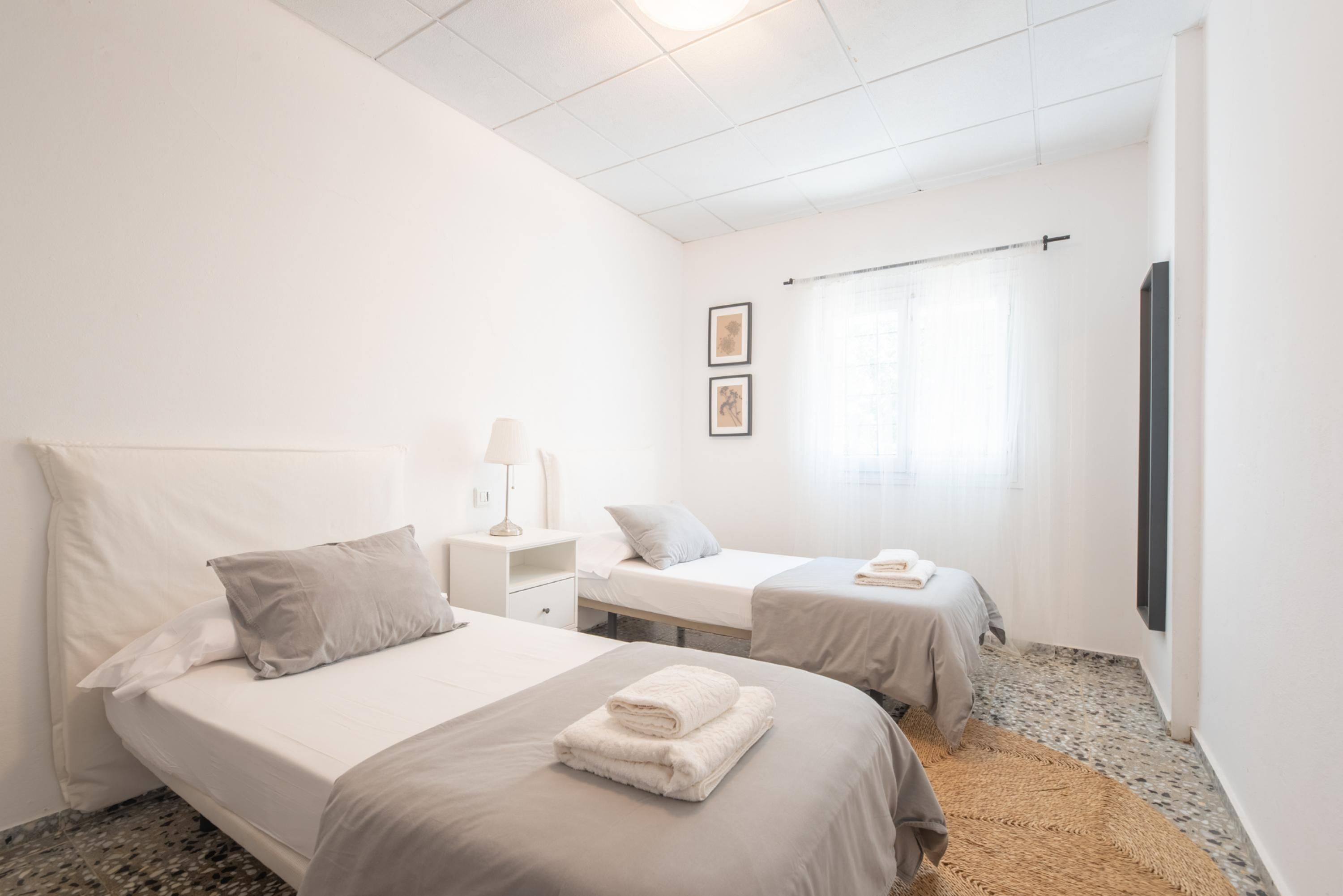 A hotel room with two beds, a small bedside table with a lamp, a wall-mounted TV, and a window with curtains.