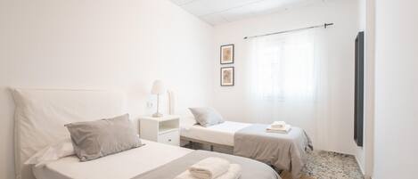 A hotel room with two beds, a small bedside table with a lamp, a wall-mounted TV, and a window with curtains.