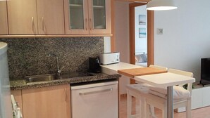 Una cocina compacta con encimeras de granito, lavavajillas integrado, una pequeña mesa de comedor con una silla y un televisor de pantalla plana.