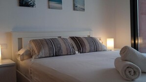 Una cama bien hecha con almohadas a rayas y toallas enrolladas, una lámpara de noche y una ventana con vista a un edificio rojo.