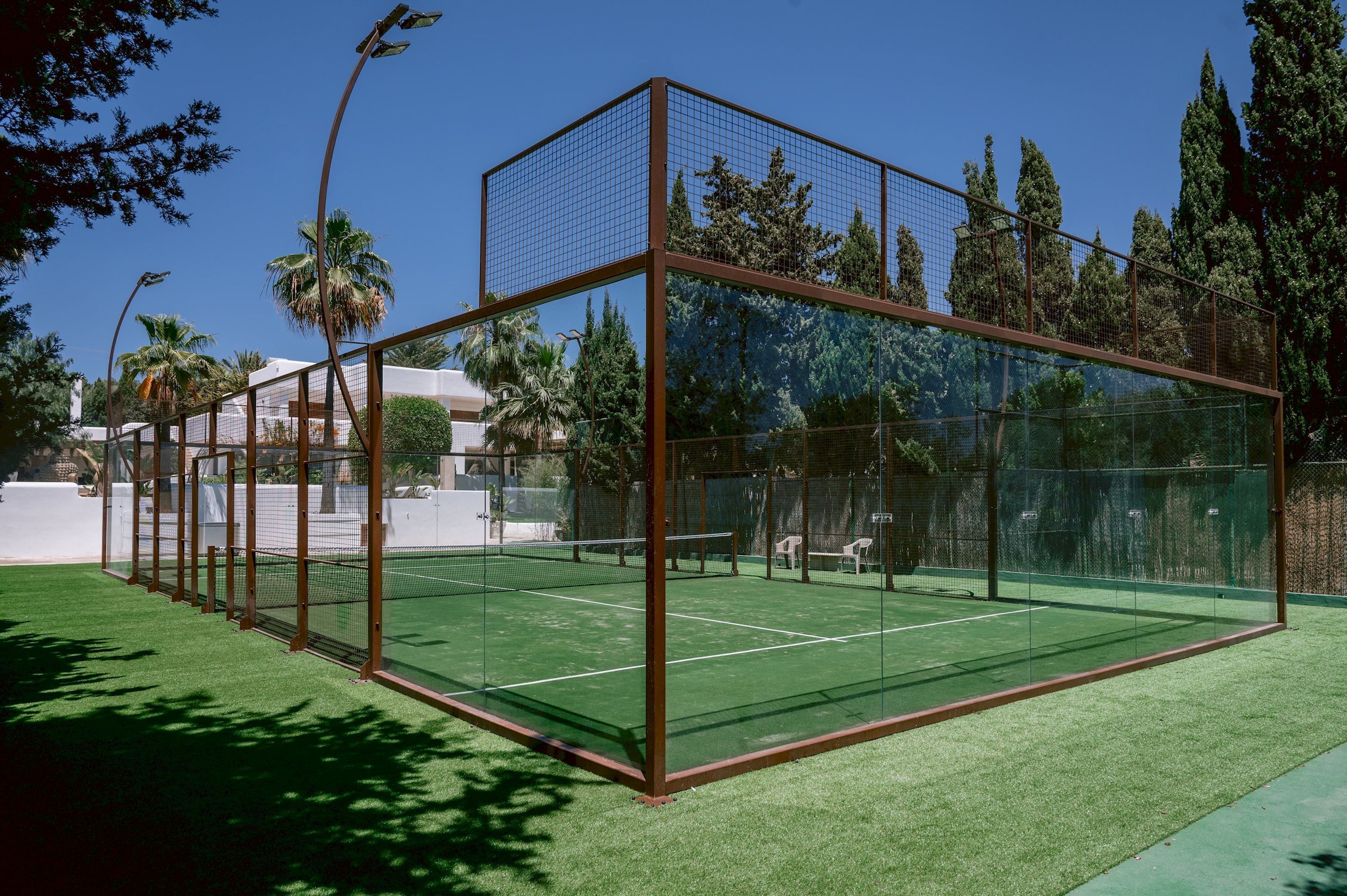 A tennis court with a glass enclosure, surrounded by trees and a white wall.