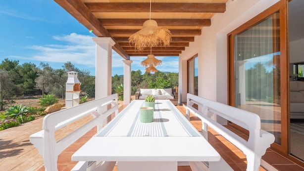 A covered outdoor patio with a white table and benches, a hanging light fixture, and a view of the surrounding landscape. - Villa Sa Planeta in Sant Josep de Sa Talaia (Sant Josep de sa Talaia)