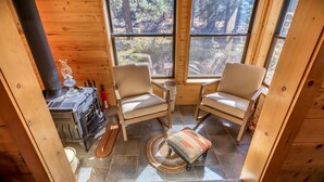 Acogedor interior de cabaña con dos sillones, una mesita, chimenea y vistas al bosque a través de amplios ventanales.