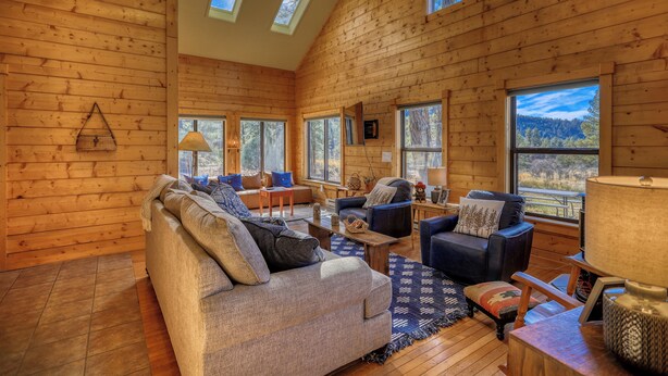 Interior acogedor de una cabaña de madera con sofá, sillas y una mesa de comedor. Amplias ventanas ofrecen vistas del exterior.