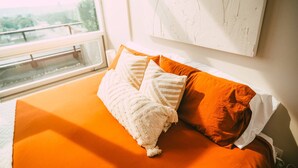 Un lit avec une literie orange et des oreillers blancs, une fenêtre donnant sur un paysage agréable et un tableau abstrait blanc accroché au mur.