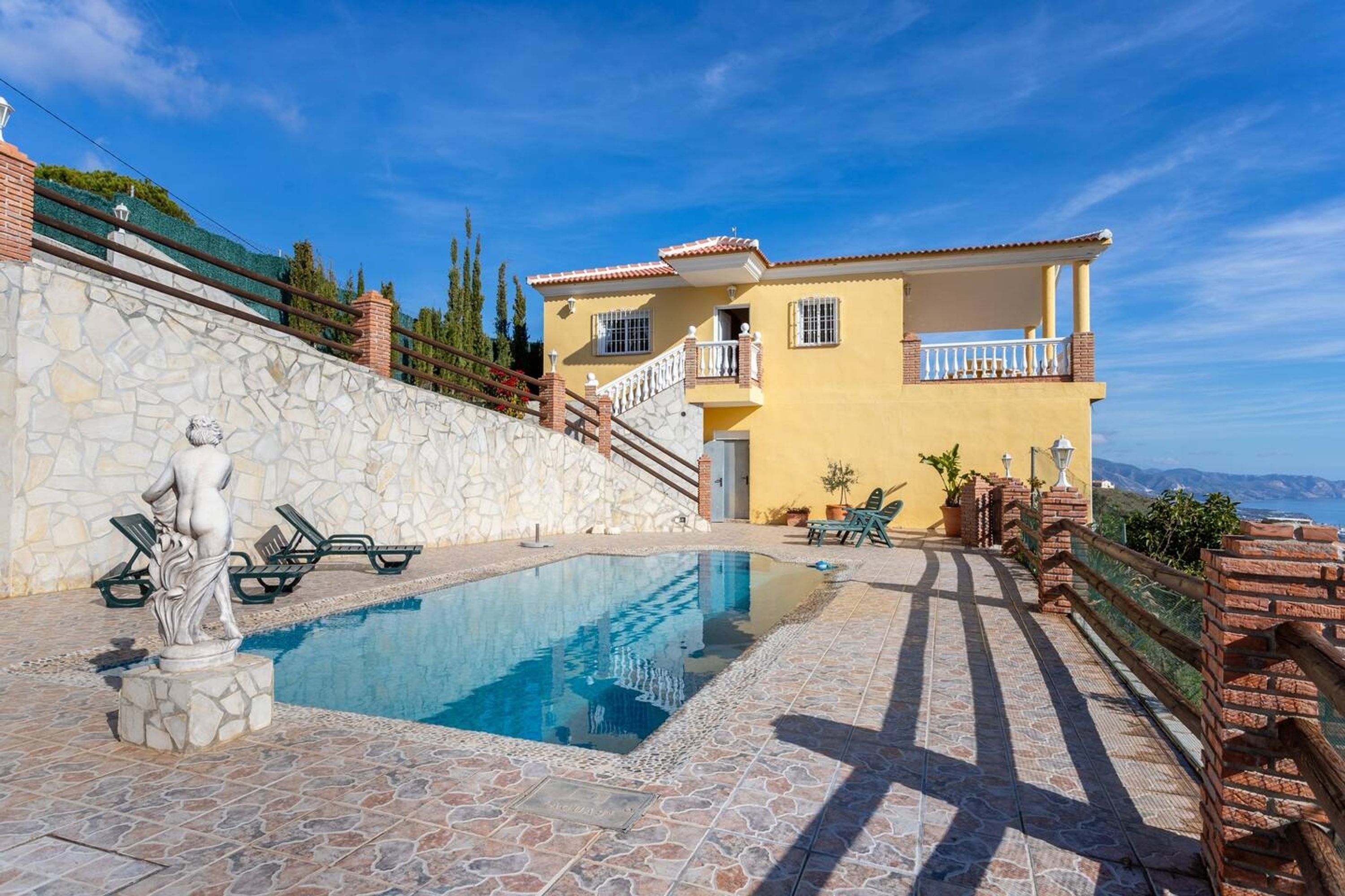 A pool area with a stone wall, a statue, and a yellow building with a balcony.