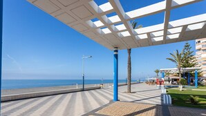 A seaside promenade with a clear view of the ocean, a covered walkway, and a building with blue pillars. - Apartamento Torrox Playa Solset (Torrox)