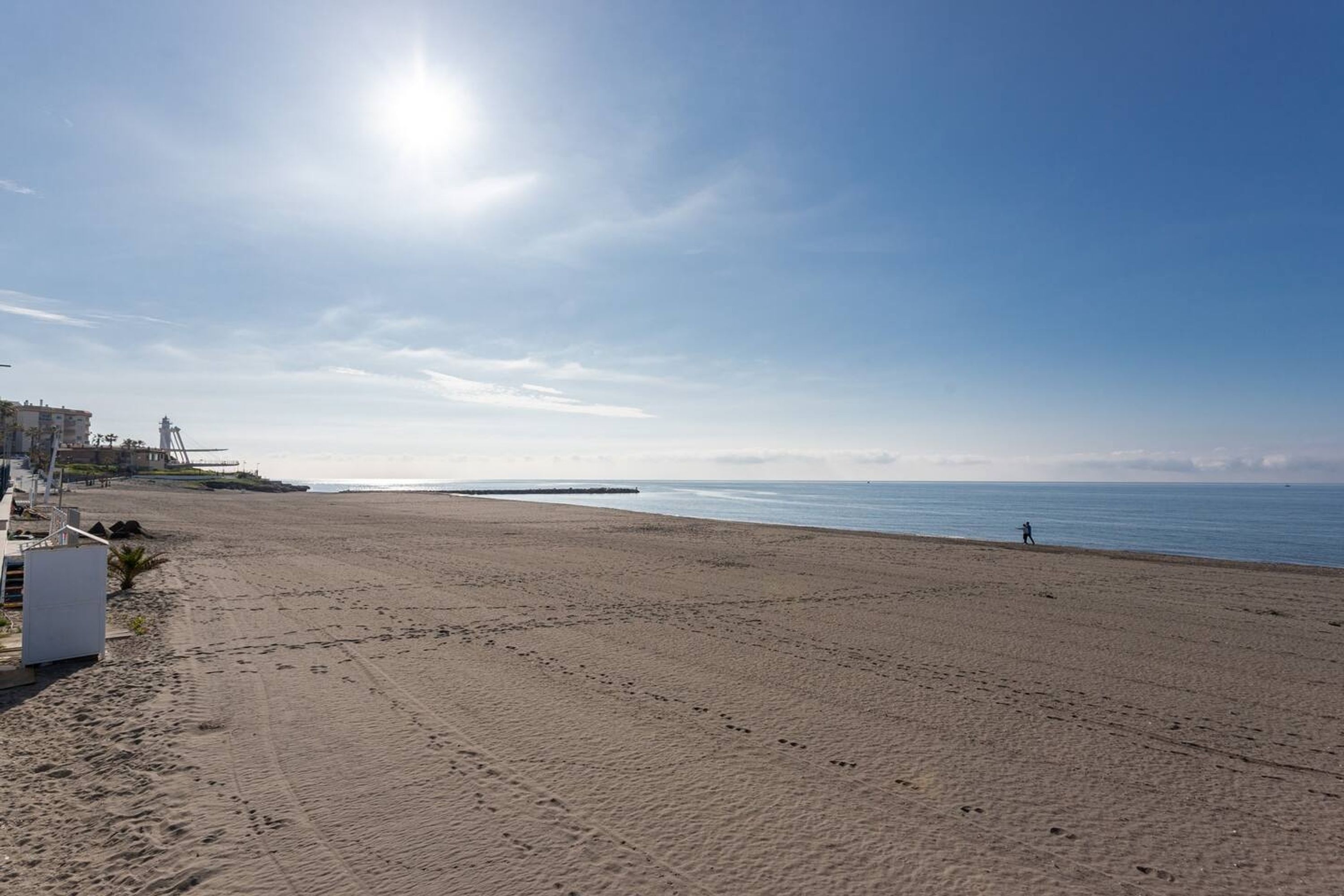 Een breed zandstrand met voetafdrukken, een pier en een persoon die loopt.