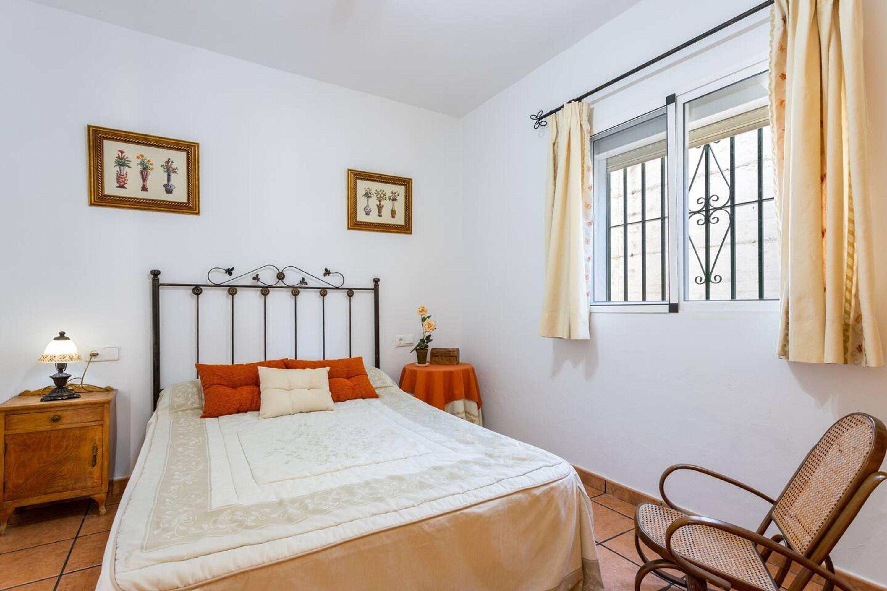 Une chambre à coucher comprenant un lit, une table de chevet, une chaise, une fenêtre avec des rideaux et deux tableaux encadrés accrochés au mur.