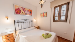 Un dormitorio con cama, mesita de noche de madera, luz empotrada en la pared y ventana con contraventanas de madera.