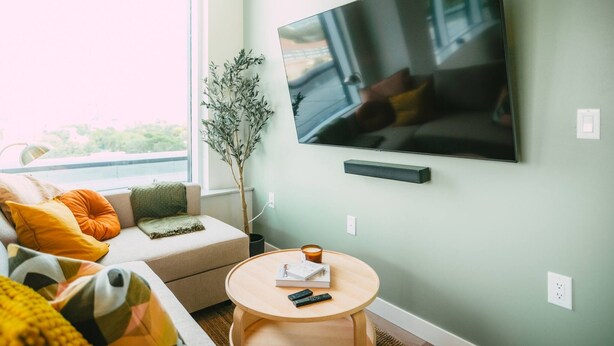 A modern living room with a flat-screen TV mounted on the wall, a sofa with colorful cushions, a small round table with a remote control, and a potted plant. - 903 (Halifax)