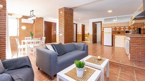 A modern living room with a grey sofa, a dining area with white chairs, and a kitchen with a brick wall and white cabinets. - Apartamento Alberti Solset (Frigiliana)