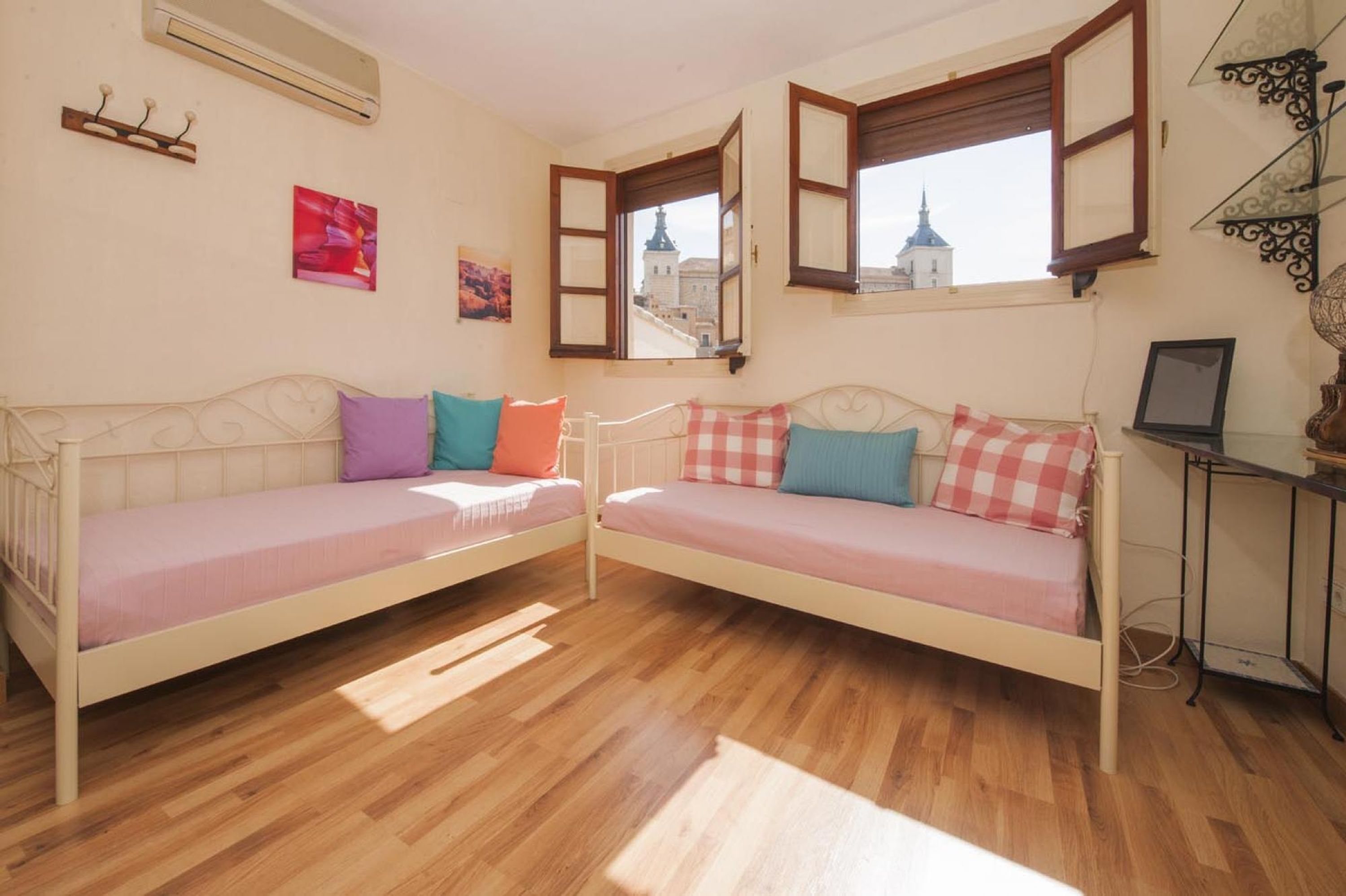 A room with two beds, wooden flooring, a window with an open shutter, and a small table with a lamp.