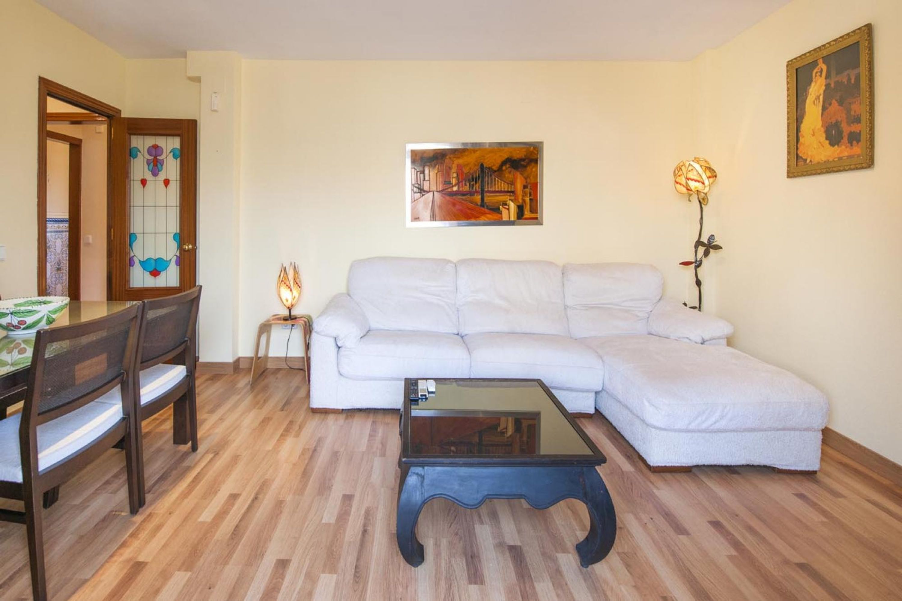 Un salón con sofá blanco, una mesa de centro de vidrio, suelo de madera, una mesa de comedor con sillas, una puerta de vitral y dos lámparas de pared.