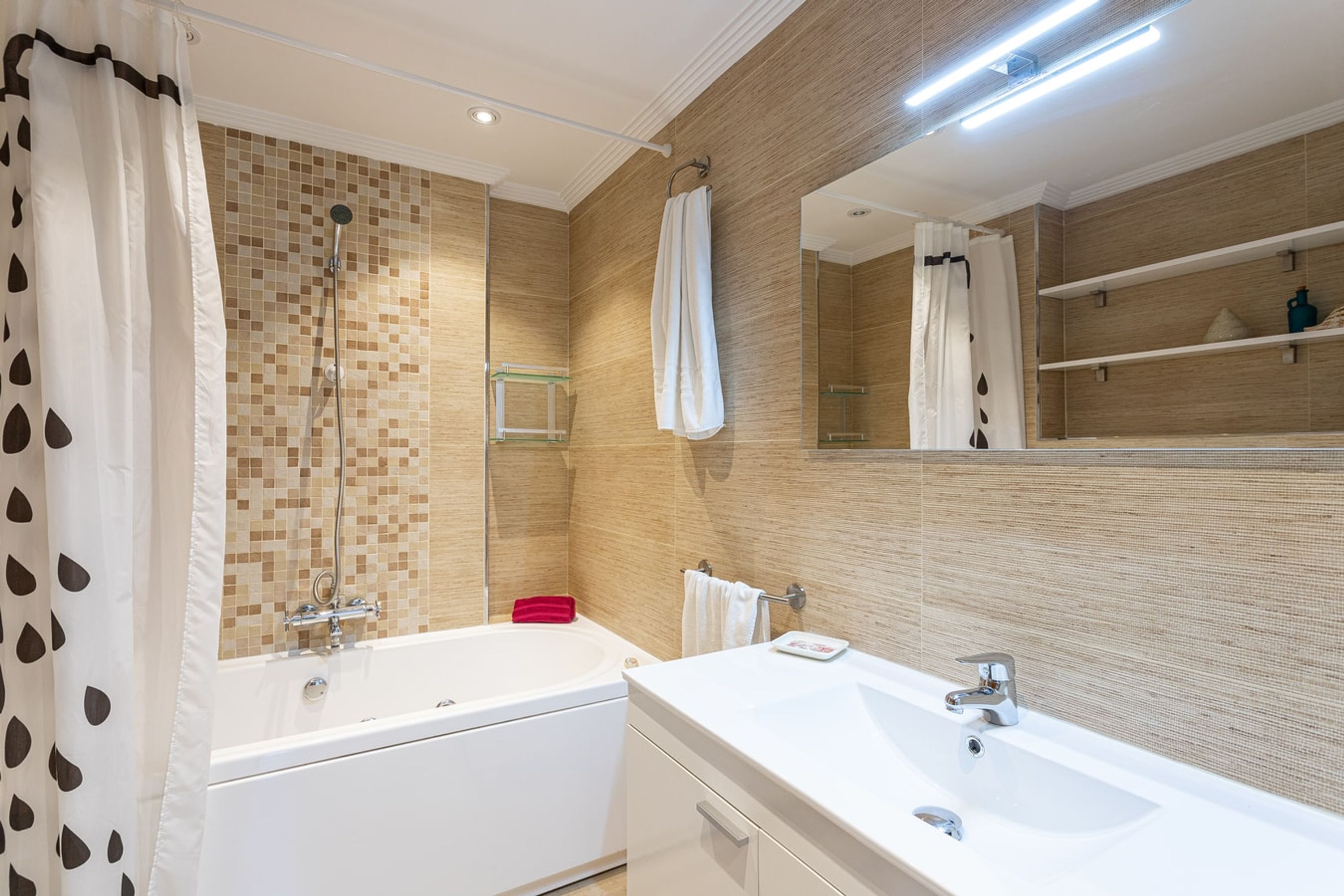 Ein modernes Badezimmer mit Badewanne, Waschbecken und Dusche. Es gibt Ablagen mit Pflegeprodukten und einen Handtuchhalter.