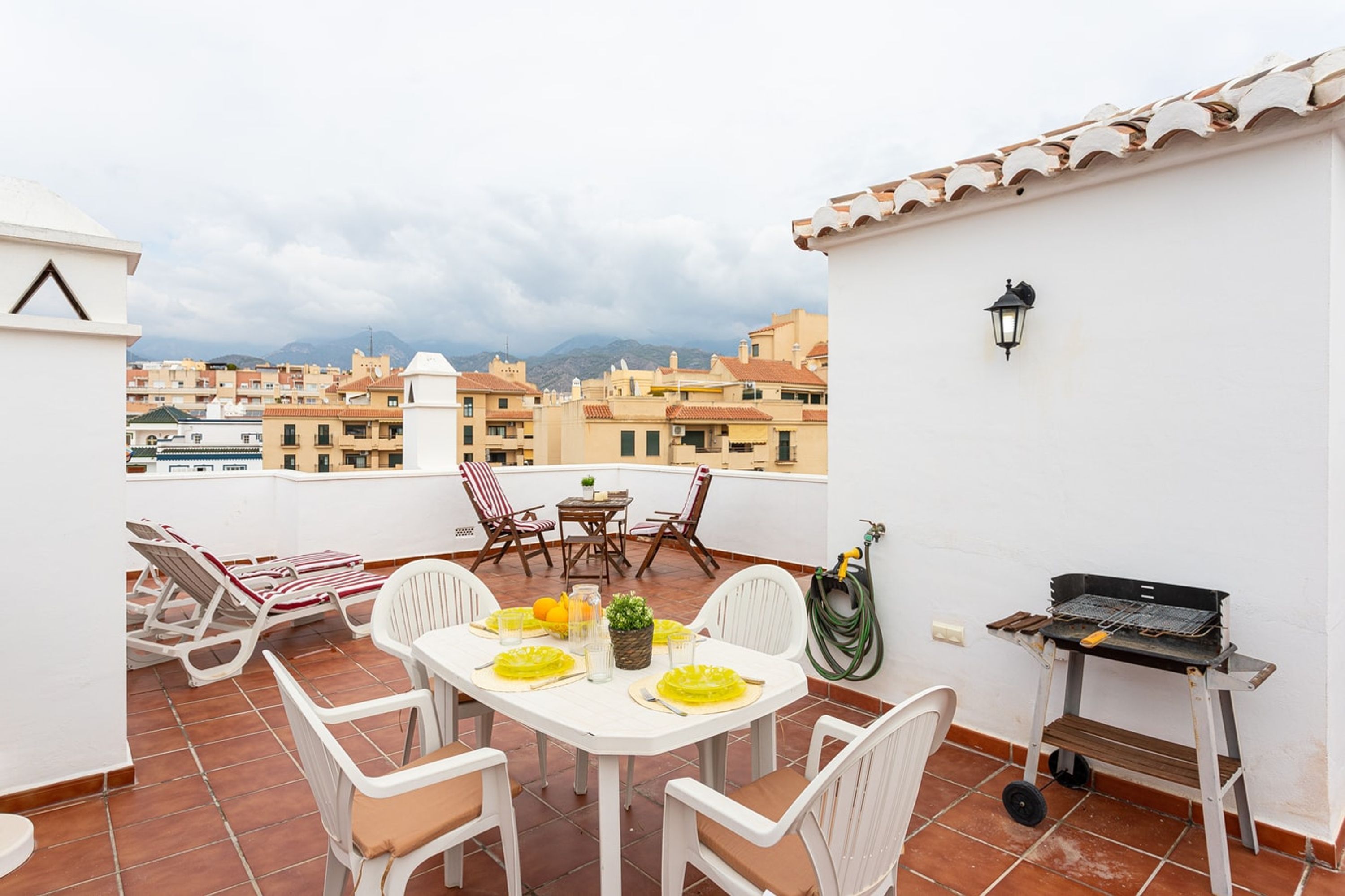 A rooftop terrace with a table set for dining, outdoor chairs, a grill, and a view of the cityscape.