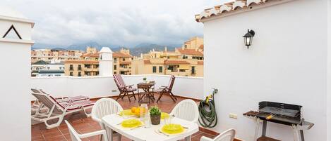 A rooftop terrace with a table set for dining, outdoor chairs, a grill, and a view of the cityscape.