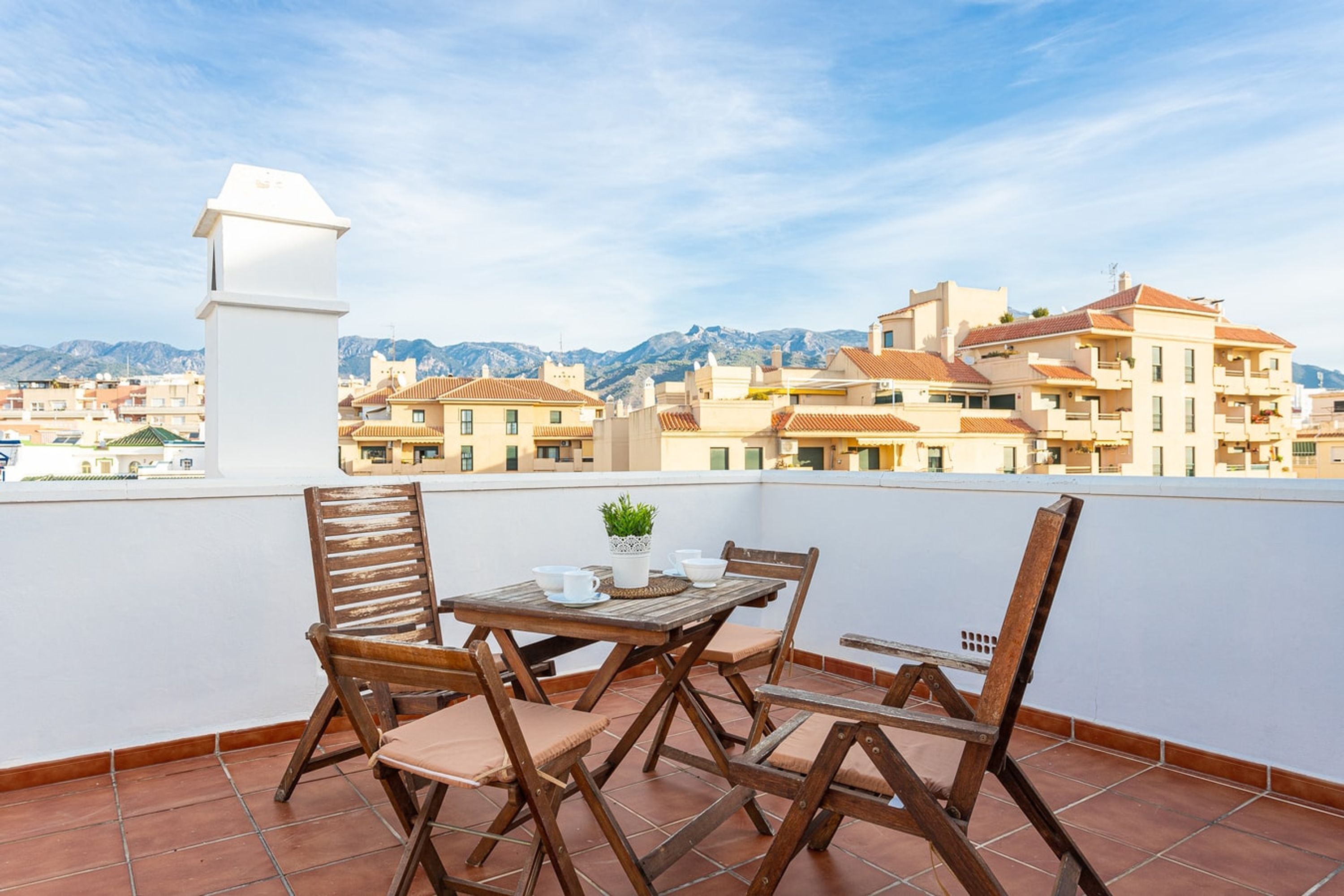 A terrace with a table and chairs, a potted plant, and a view of buildings and mountains in the background.
