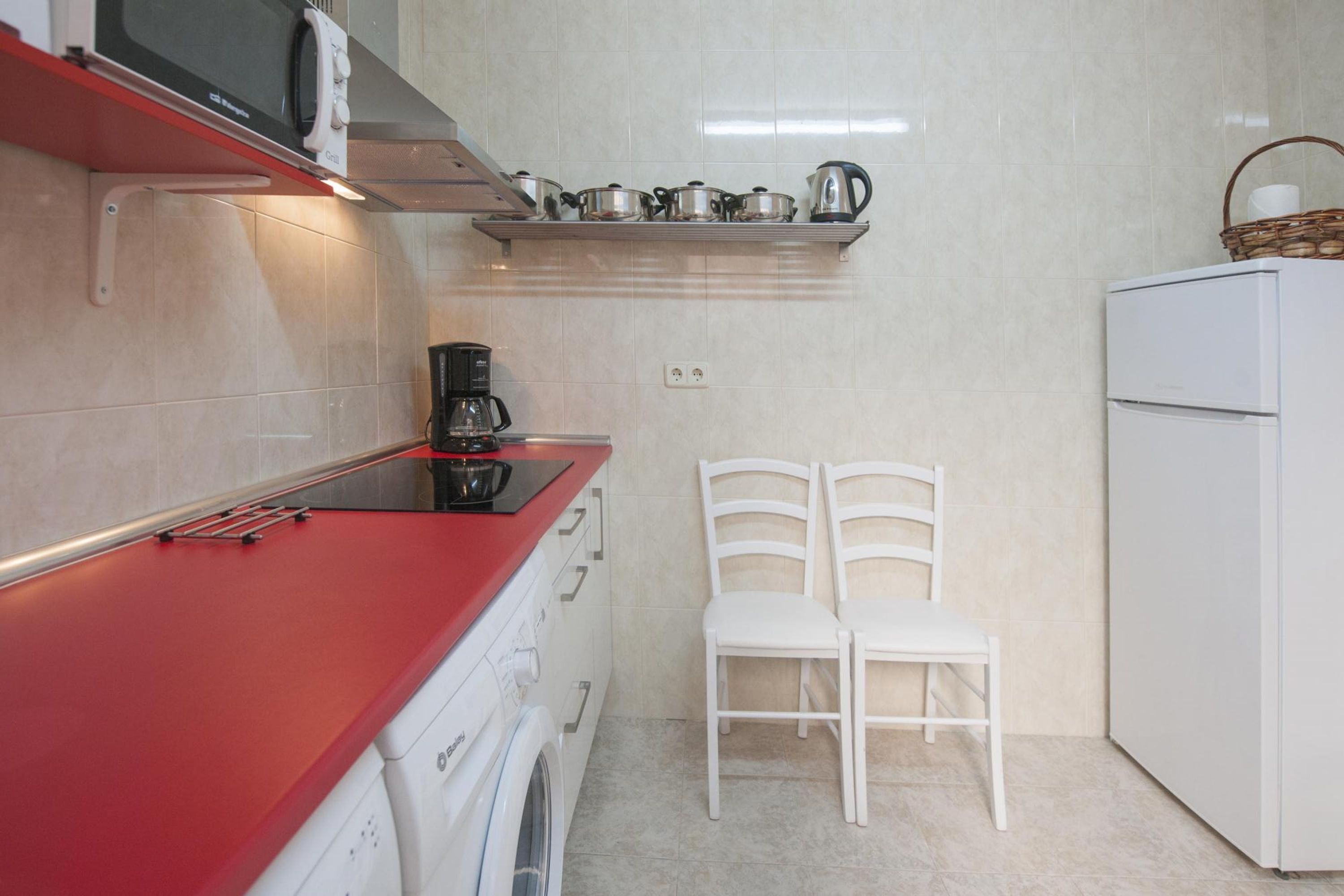 Een kleine keuken met een rood aanrecht, een koffiezetapparaat, een magnetron, een waterkoker en een witte koelkast. Er staan twee witte stoelen en boven het aanrecht bevindt zich een plank met keukengerei.
