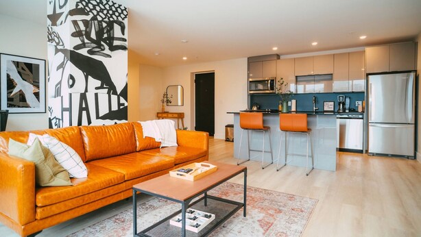 A modern living room with a sofa, coffee table, and a kitchen area with a refrigerator, microwave, and bar stools. - 701 (Halifax)
