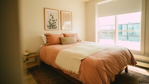 A bedroom with a large window, a bed with a beige and pink bedding, a small bedside table with a lamp, and two framed pictures on the wall. - 701 (Halifax)