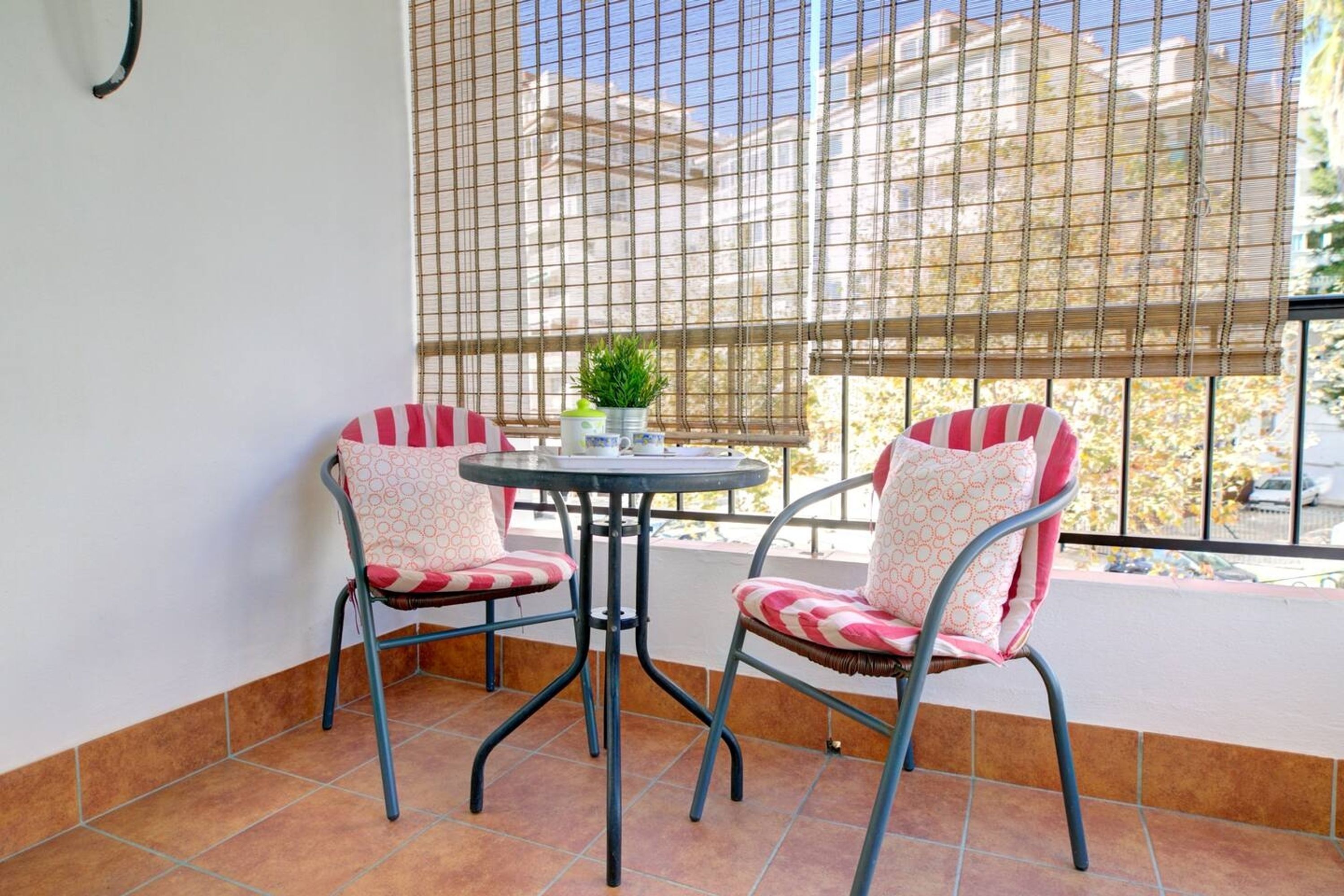 Un balcon avec deux chaises et une petite table, une plante en pot, et une vue sur un immeuble à travers une fenêtre munie d’un moustiquaire.