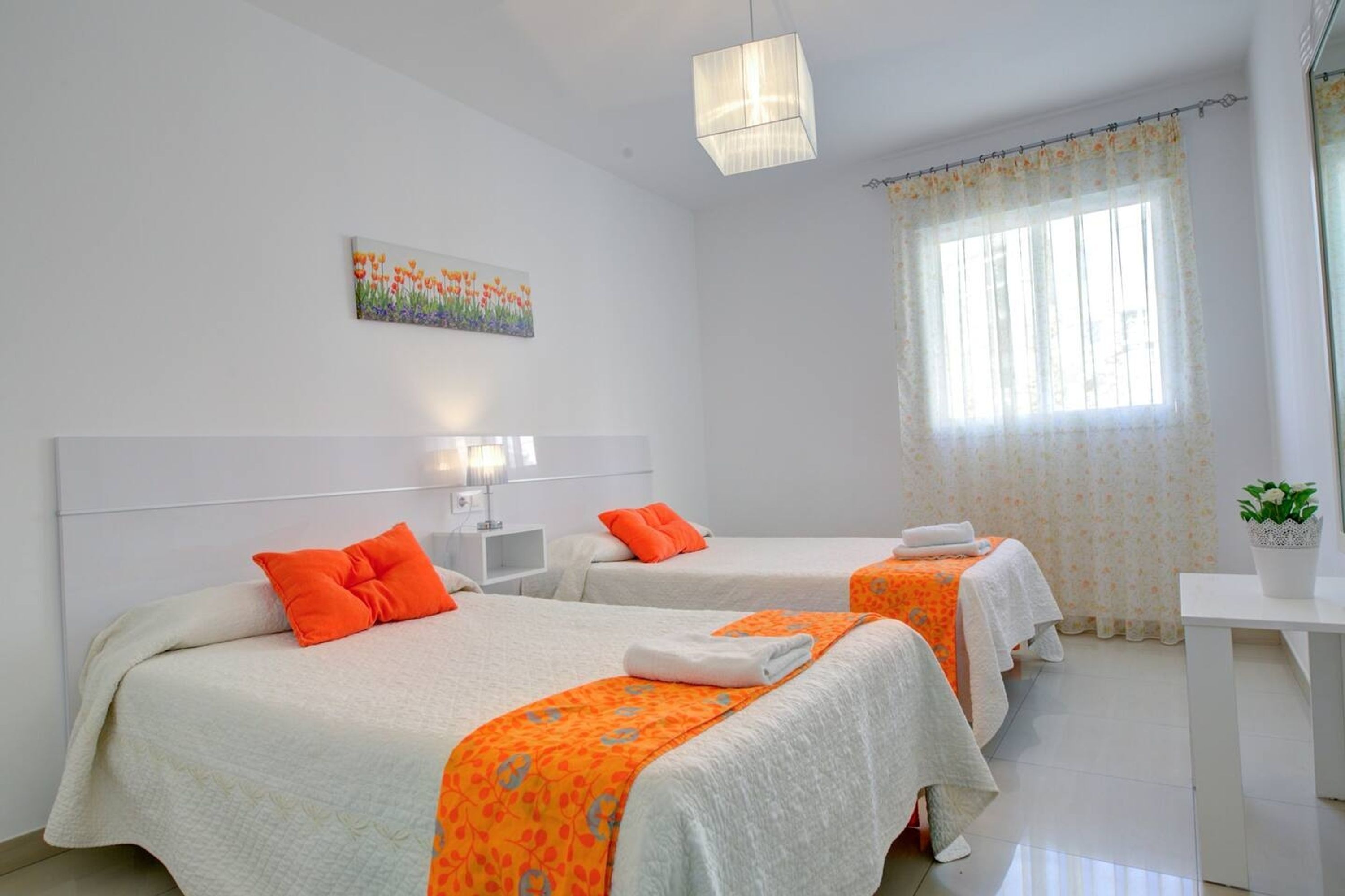 A hotel room with two beds, each with an orange blanket and pillow, a white headboard, a painting on the wall, and a window with a floral curtain.