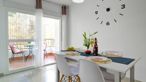 A dining area with a table set for two, a clock on the wall, and a balcony with chairs and a table. - Apartamento Palmeras 29 Solset (Nerja)