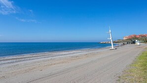Una playa con una torre de salvavidas blanca, cielo azul despejado y edificios al fondo.