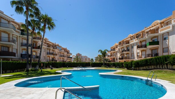 Una piscina rodeada de edificios de apartamentos con balcones, un cielo azul despejado y abundante vegetación bien cuidada.