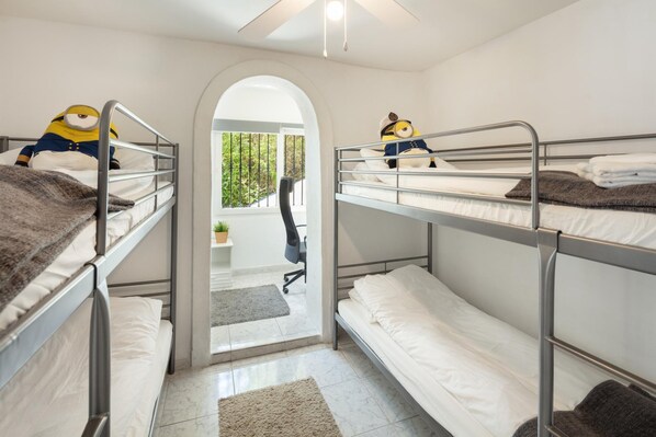 A bunk room with three bunk beds, each with a Minion plush toy on top. There is a desk and chair in the center, and a small potted plant on the desk.