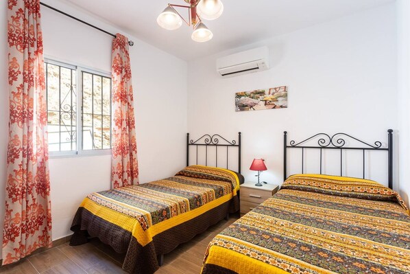 Habitación con dos camas, cada una con ropa de cama estampada, una ventana con cortinas, una lámpara de noche y un equipo de aire acondicionado en la pared.