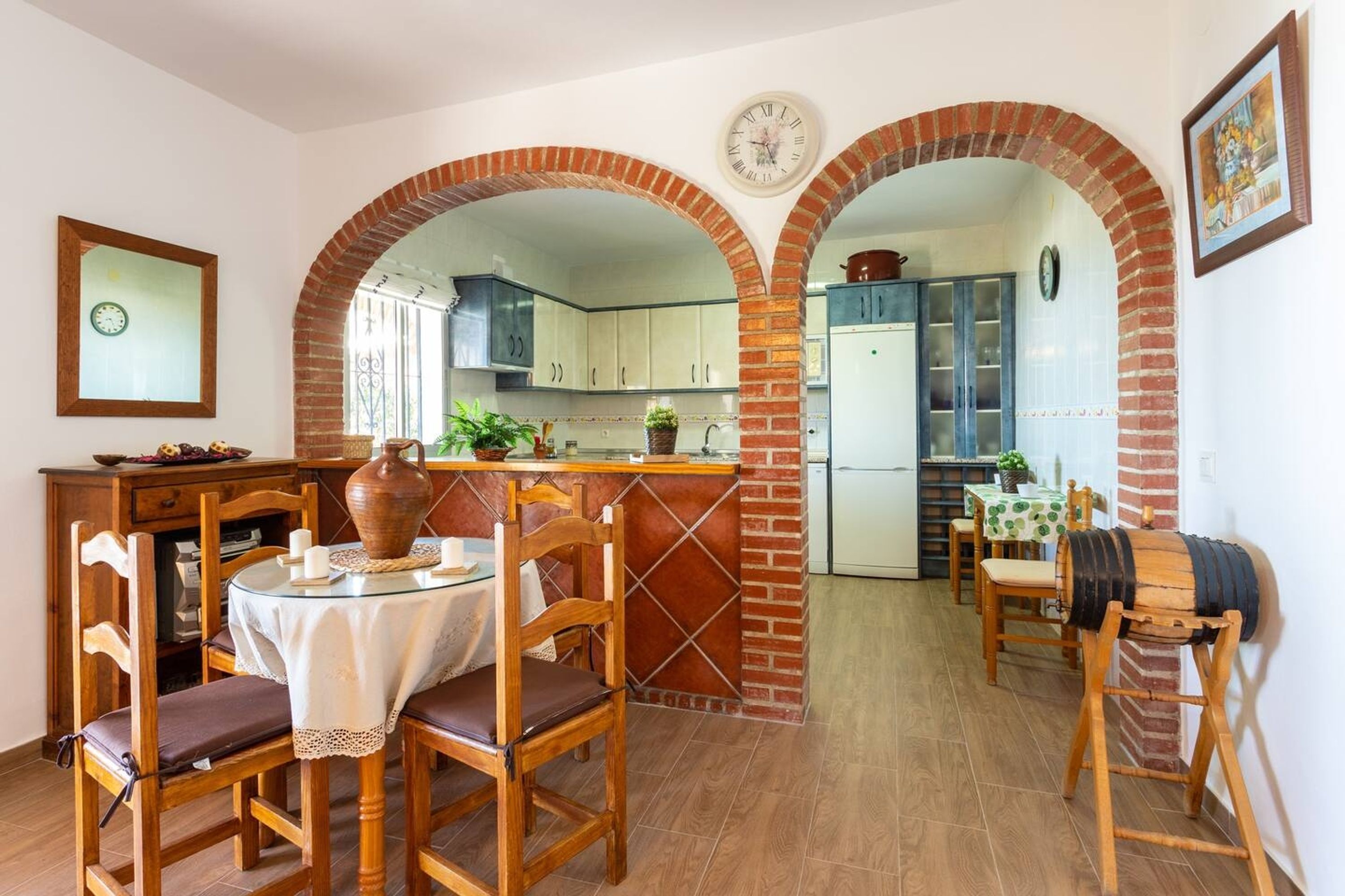 A dining area with a round table, chairs, and a rustic brick archway leading to a kitchen.