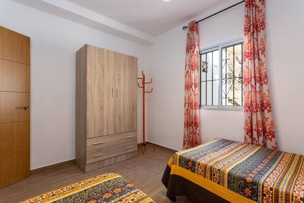 Une chambre à coucher avec une armoire, un porte-manteau, un lit recouvert d’une couvre-lit à motifs et une fenêtre avec des rideaux.