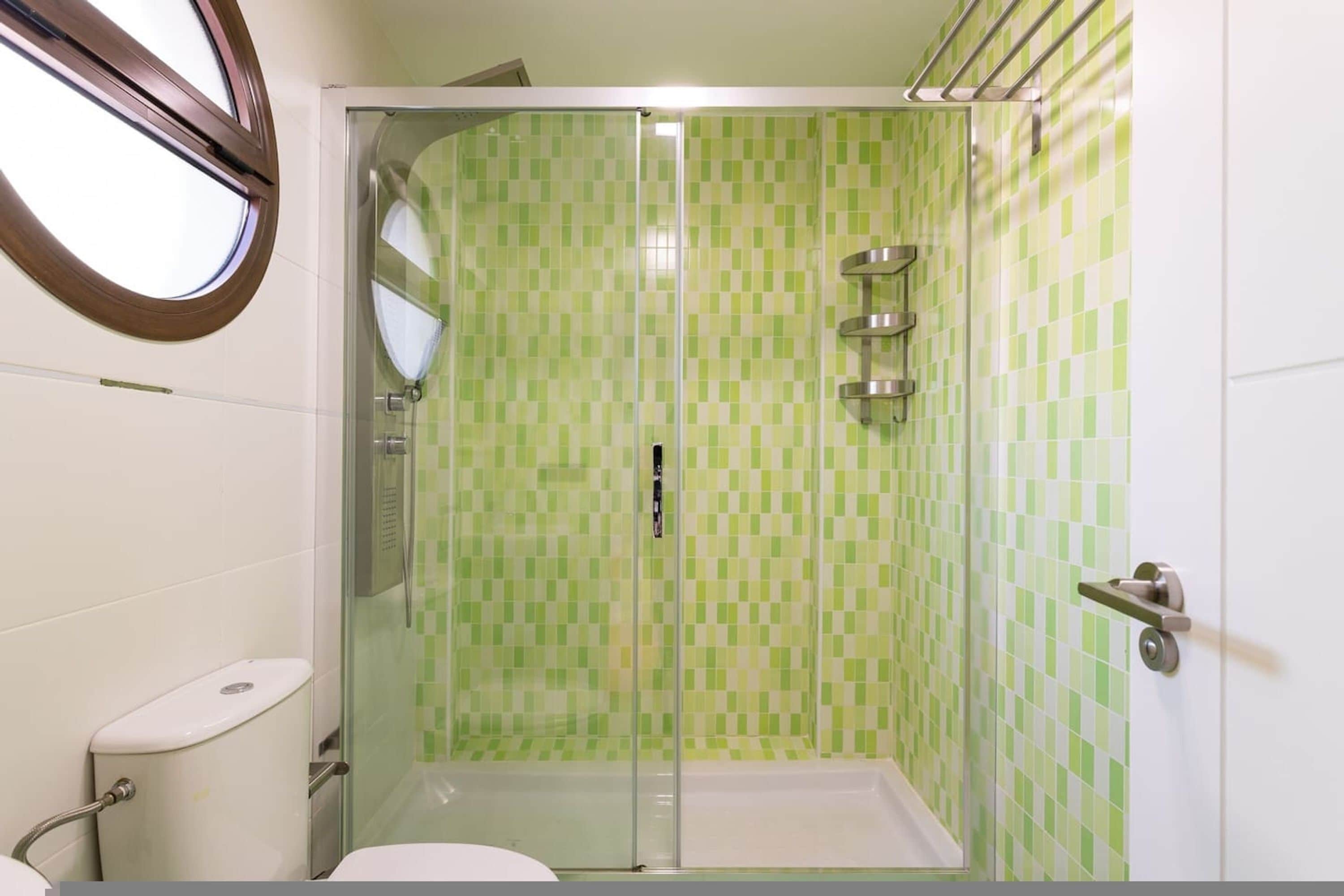 A bathroom with a glass-enclosed shower, green checkered tiles, a toilet, and a window.